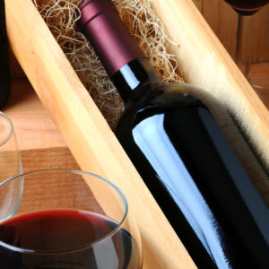 A red wine bottle in a wooden box filled with straw on a tasting room table with two glasses of poured wine.