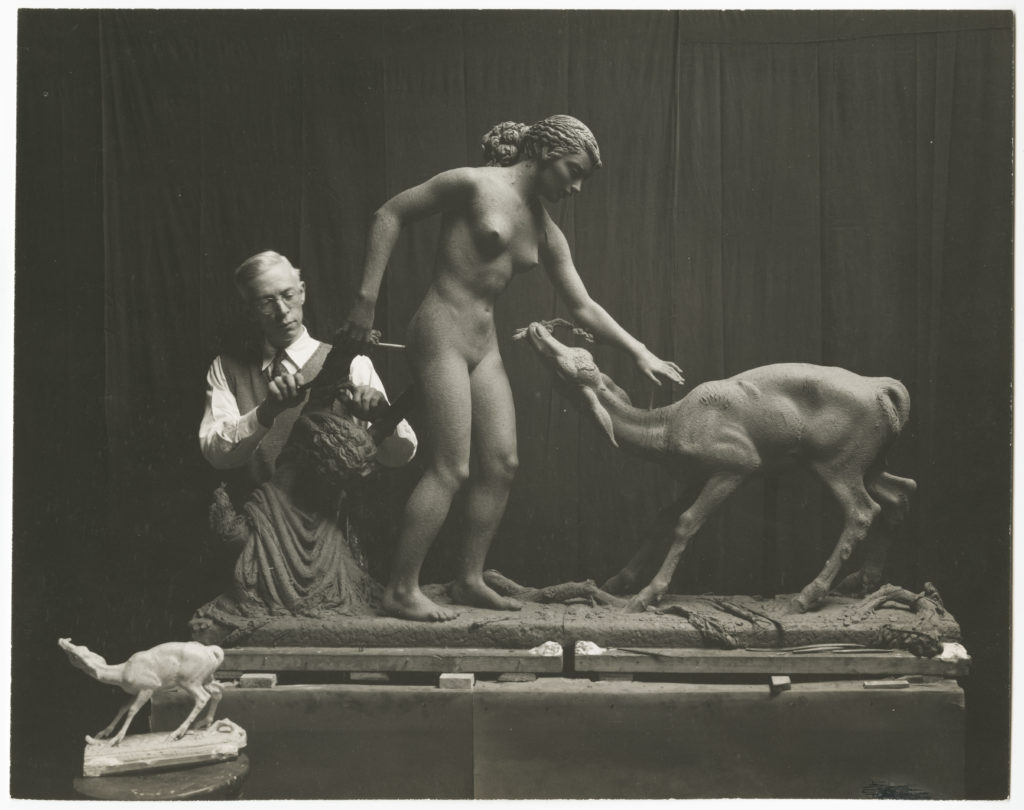 Black and white photo - a man works on a sculpture of a woman and a deer